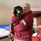 Smiling man in red hoodie holding a black dog in a red sweater in a living room